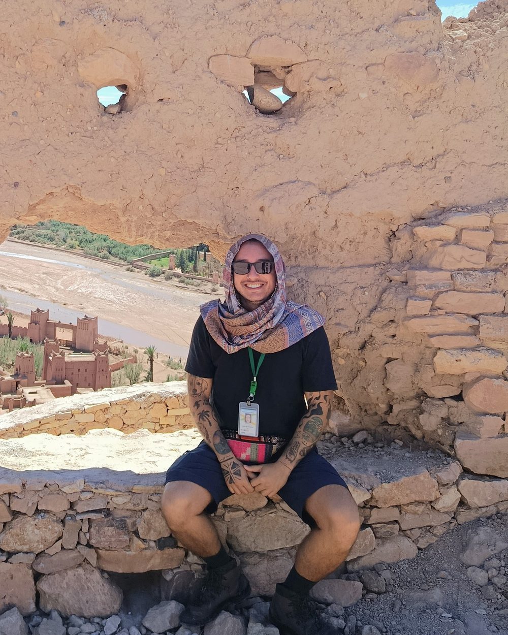 Ricardo Cardoso sorrindo, com lenço na cabeça e crachá de guia, sentado em uma muralha de barro no Marrocos